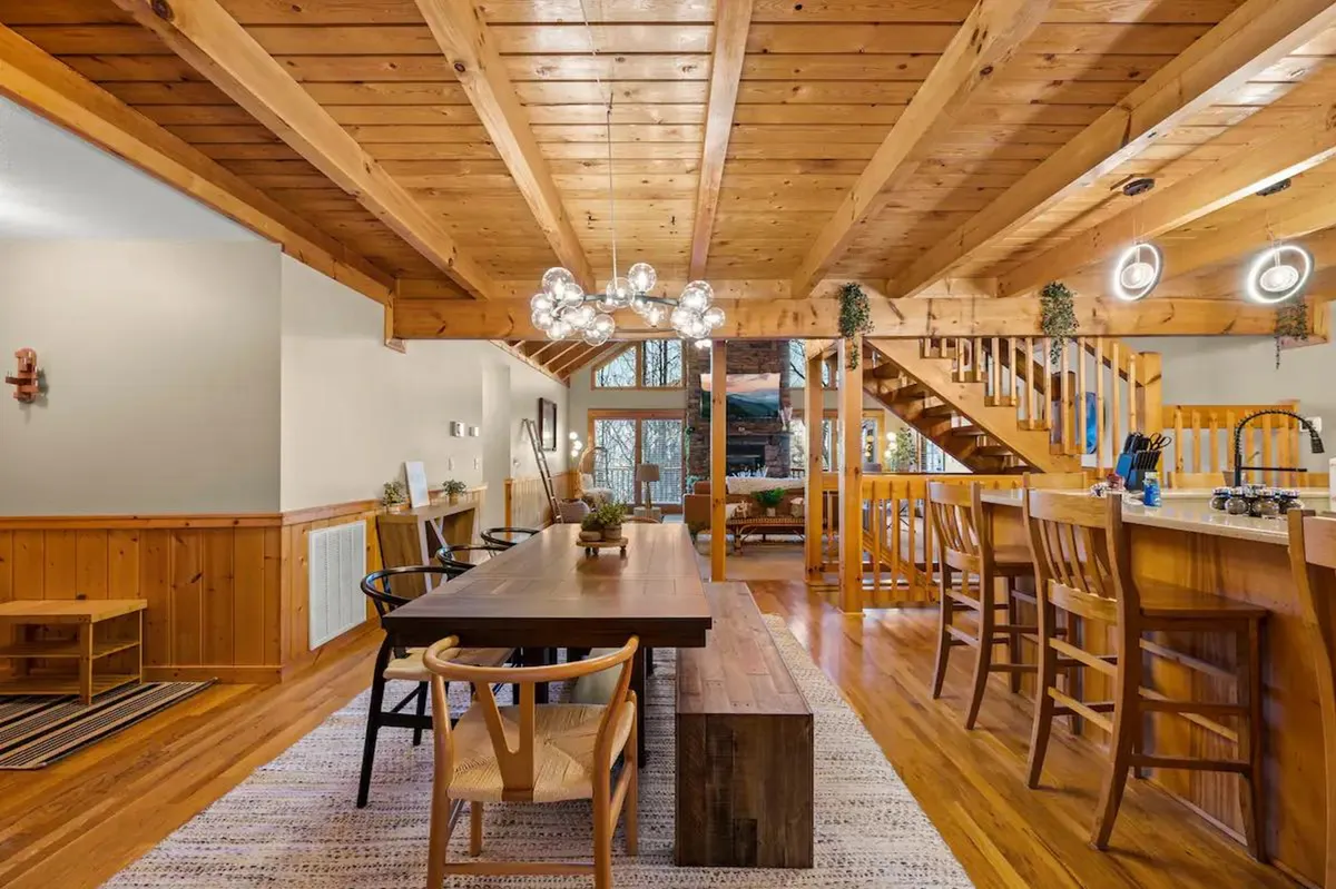 A spacious interior view featuring a long wooden dining table set beneath an elegant chandelier, with an open staircase and a visible bar area nearby. The scene radiates warmth and hospitality, set up for shared meals and festive gatherings.