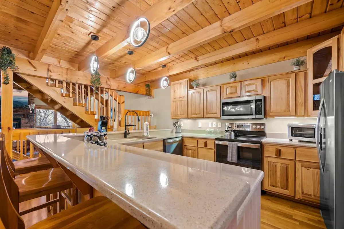 This image showcases a warm, modern kitchen with an island counter, sleek wooden cabinetry, open staircase, and stylish pendant light fixtures. The kitchen is set up for both cooking and gathering, with visible counter seating that invites interaction and shared moments.
