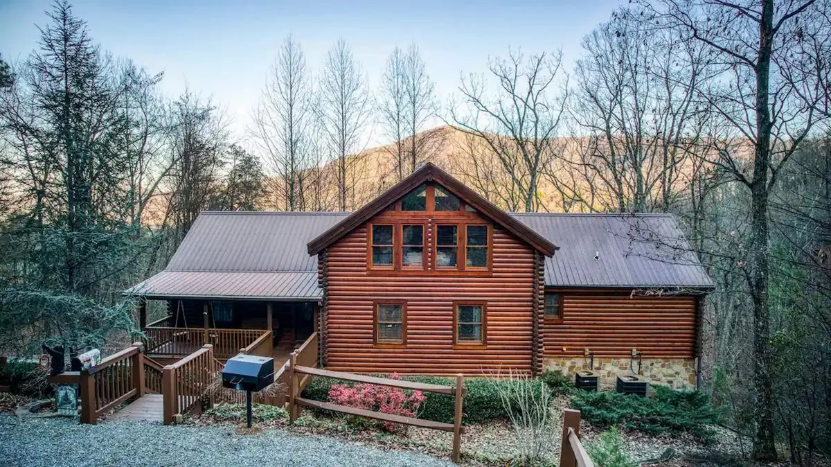 The image shows a stunning log cabin nestled amidst lush trees with a mountain backdrop. The spacious front porch invites relaxation, and the landscaped garden hints at outdoor gatherings. Warm sunset light bathes the cabin, highlighting its rustic charm and serene setting.