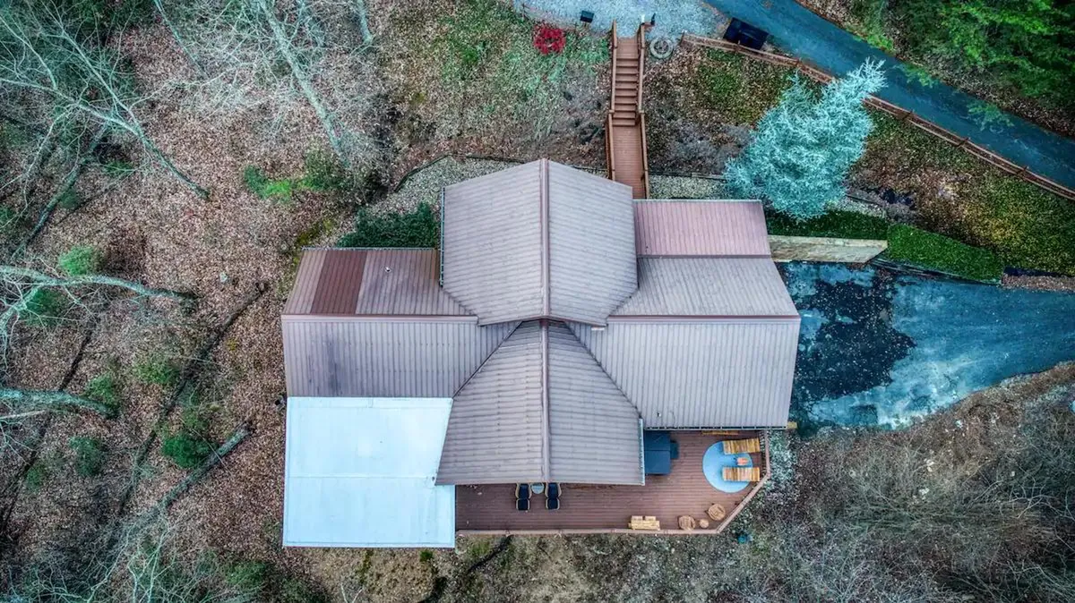 A peaceful exterior view of the cabin, showing its distinctive roofline, a welcoming deck area, and a pathway leading through surrounding trees. The deck is set up for relaxation and blends seamlessly with the natural landscape, inviting guests to step outside and explore.