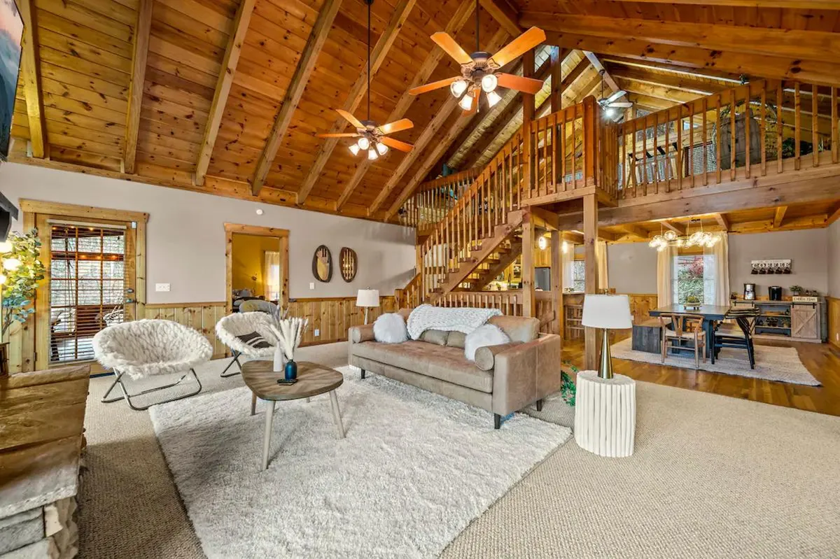 This angle looks across an open-plan living and dining space, capturing wooden beams, a comfortable sofa, stylish armchairs, a visible dining area, and a staircase. The cozy yet spacious layout encourages relaxation and socialization for groups.