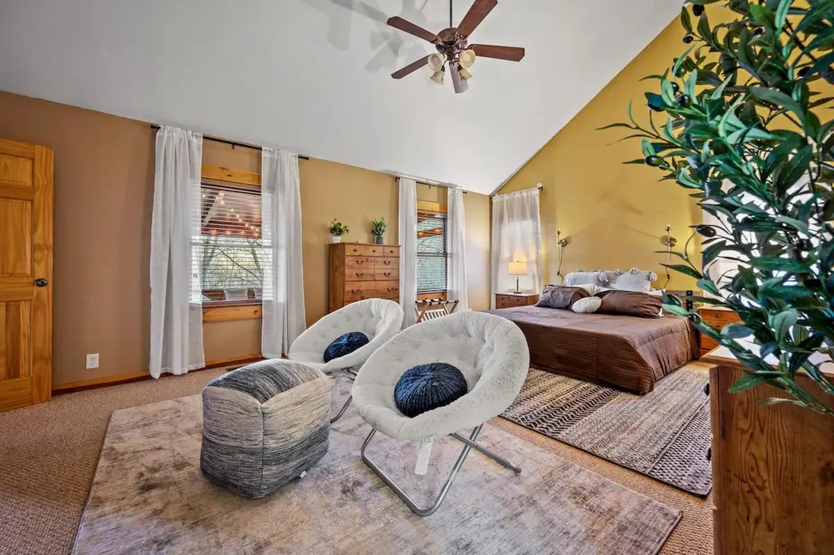 A cozy bedroom featuring a comfortable bed with soft bedding, a small seating area, a dresser, and potted plants. Soft natural light fills the room through windows, creating a tranquil and restful atmosphere.