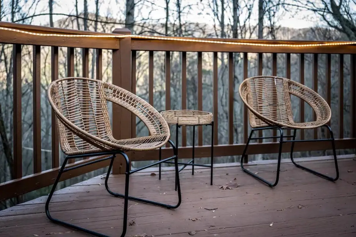 A cozy section of the cabin’s wooden deck featuring two wicker chairs and a small side table, framed by a rustic railing. The setting is ideal for relaxing, socializing, or simply soaking in the tranquil forested surroundings.