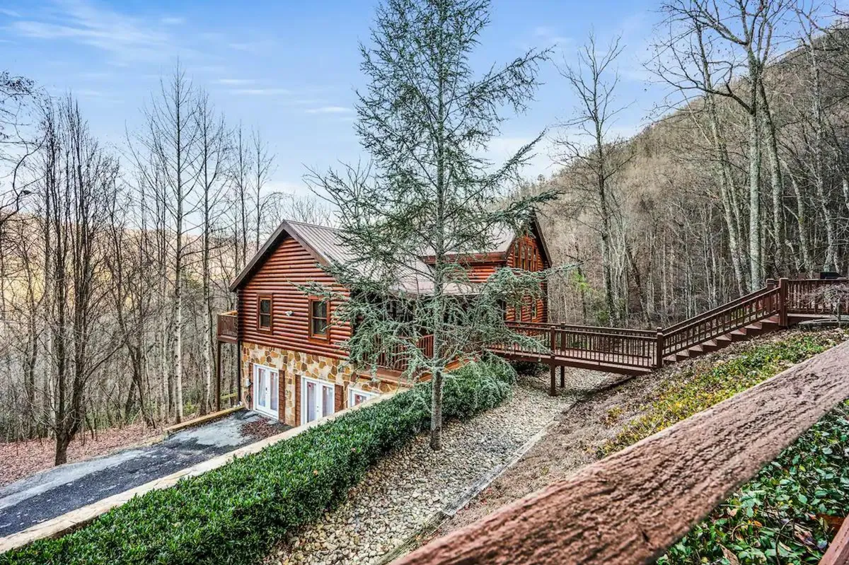 A serene exterior view of a log cabin nestled among trees, featuring a spacious deck area that invites guests to relax and enjoy the natural surroundings. The scene captures the inviting wooden structure, lush greenery, and the tranquil atmosphere perfect for outdoor activities or unwinding after a day of adventure.
