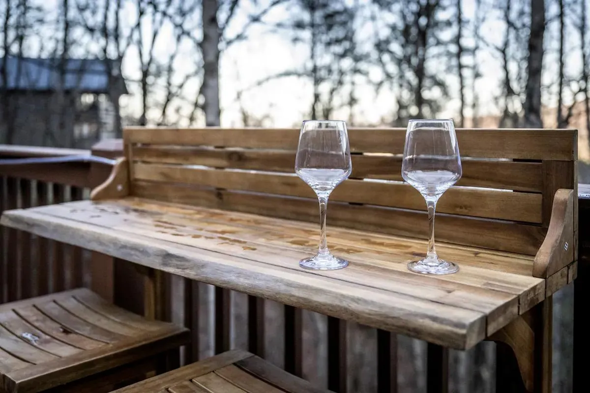 A serene outdoor wooden bar setup with wine glasses arranged on the counter, surrounded by a lush natural backdrop. The scene captures a small group enjoying drinks outdoors, suggesting relaxation and celebration as the sun sets.