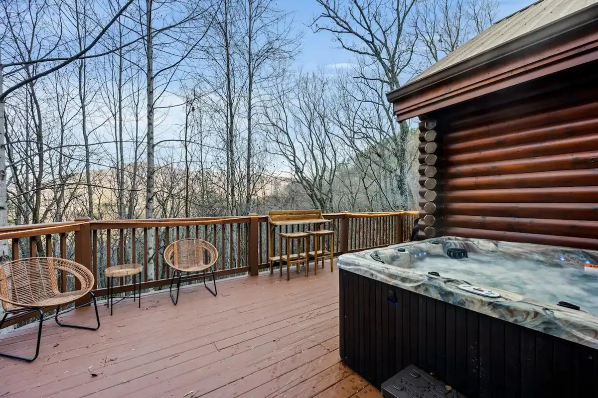 A tranquil view of a hot tub nestled on a wooden deck surrounded by trees, with comfortable outdoor seating for relaxing and taking in the natural beauty. The ambiance is peaceful and inviting, ideal for unwinding after a day out.