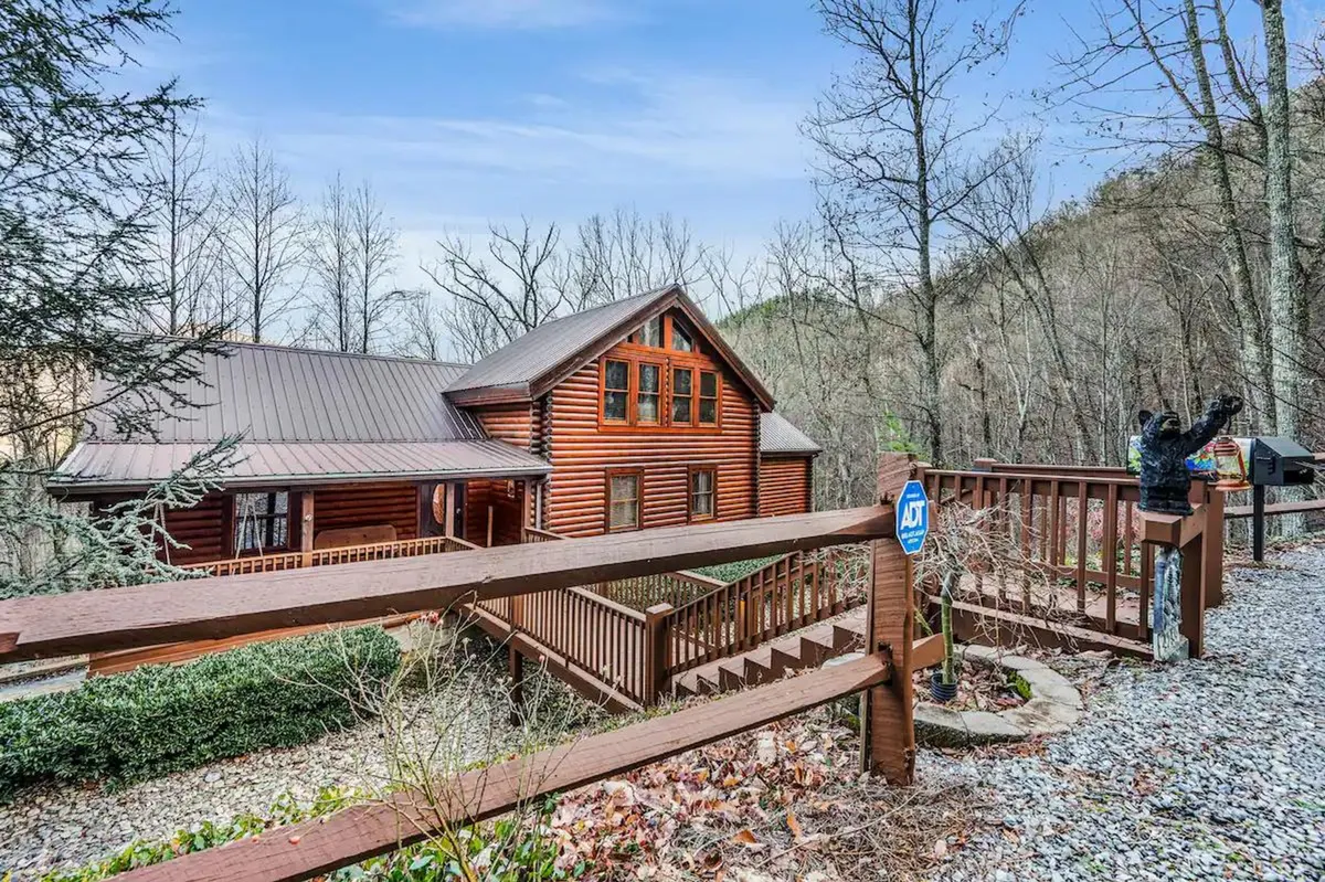 A welcoming view of a log cabin exterior, highlighting a wooden deck and its integration with the surrounding forest. A staircase leads to the entrance, and the scene conveys a sense of arrival, adventure, and possibility for group gatherings and outdoor experiences.